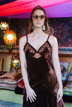 Woman wearing a brown velvet boho dress with floral details in a colorful, possibly Middle Eastern, setting.