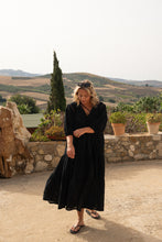 Woman in a black boho dress standing in a scenic outdoor setting with rolling hills and stone wall.
