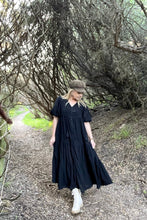 Woman in a black boho dress and white shoes walking on a path with trees in the background
