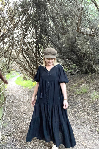 Woman in a black boho dress and cap standing on a path with trees in the background