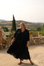 A person wearing a black midi length cotton boho dress with buttons down the front and a mandarin collar, standing in front of a scenic landscape.