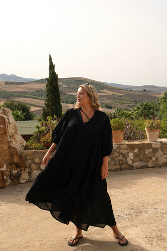 A person wearing a black midi length cotton boho dress with buttons down the front and a mandarin collar, standing in front of a scenic landscape.