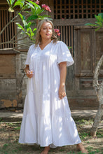 A woman wearing a white cotton knee-length boho dress with short sleeves, buttons down the front, and a mandarin collar, standing in an outdoor setting.