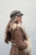 Woman wearing a brown coat with fur trim and a patterned cap against a white brick wall.
