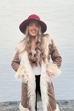 Woman wearing a brown coat, white scarf, and red hat against a light gray wall.