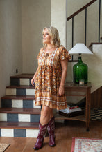 Woman in a boho patterned dress and cowboy boots standing in a home interior.
