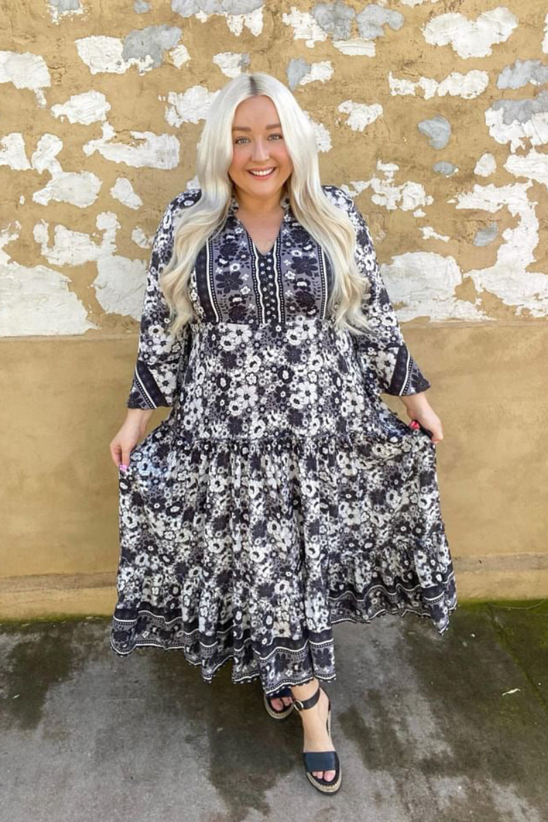 A woman standing outdoors wearing an eclectic bohemian style midi dress with a black and white floral pattern. The dress has a V-neck, 7/8 length sleeves, and a flowing skirt with a frill at the bottom.