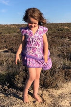 A young girl standing in a natural outdoor setting wearing a short-sleeved floral print dress in assorted colors.
